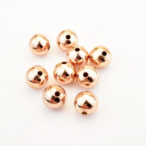 Peut inclure: Dix perles de couleur or rose, chacune avec un petit trou au centre. Les perles sont &eacute;parpill&eacute;es sur une surface blanche.