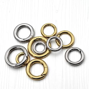 May include: A collection of circular spring gate rings in various sizes, featuring a mix of polished silver-tone and gold-tone metal finishes. Each ring includes a functional spring-loaded opening mechanism for attaching items securely.
