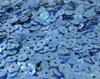 May include: A large collection of small, round, flat blue sequins. Each individual sequin features a central hole and a reflective, iridescent finish that catches the light, creating a shimmering effect across the entire pile of craft materials.