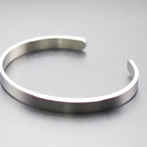 K&ouml;nnte beinhalten: Ein silberfarbenes Armreif-Armband. Das offene, minimalistische Design hat ein flaches, poliertes Band. Das Armband aus gl&auml;nzendem Metallmaterial ist vor einem dunklen Hintergrund platziert.