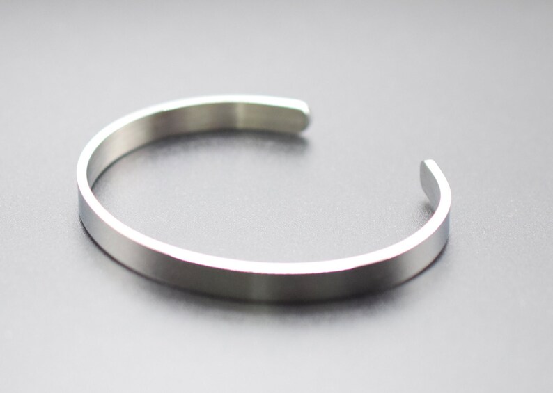 K&ouml;nnte beinhalten: Ein silberfarbenes Armreif-Armband. Das offene, minimalistische Design hat ein flaches, poliertes Band. Das Armband aus gl&auml;nzendem Metallmaterial ist vor einem dunklen Hintergrund platziert.