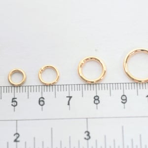 May include: A series of seven gold-toned metal jump rings arranged in increasing order of size, positioned above a ruler that shows measurements in centimeters and inches to provide a clear visual scale for each individual ring diameter.