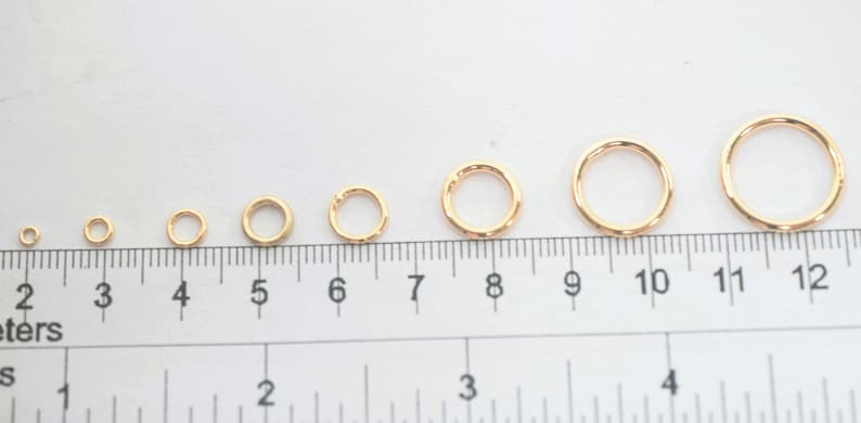 May include: A series of seven gold-toned metal jump rings arranged in increasing order of size, positioned above a ruler that shows measurements in centimeters and inches to provide a clear visual scale for each individual ring diameter.