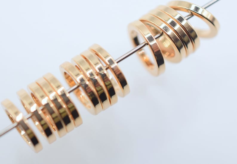 May include: A series of small, polished gold-toned metal rings with a flat, circular profile, threaded onto a thin, straight metal wire. The rings feature a smooth, reflective surface and are arranged in two distinct groups along the wire.