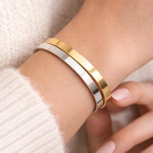 K&ouml;nnte beinhalten: Nahaufnahme von zwei minimalistischen Armb&auml;ndern, eines goldfarben und eines silberfarben. Die Armb&auml;nder werden am Handgelenk getragen, vor dem Hintergrund eines weichen, cremefarbenen Pullovers. Schlichtes, elegantes Design.