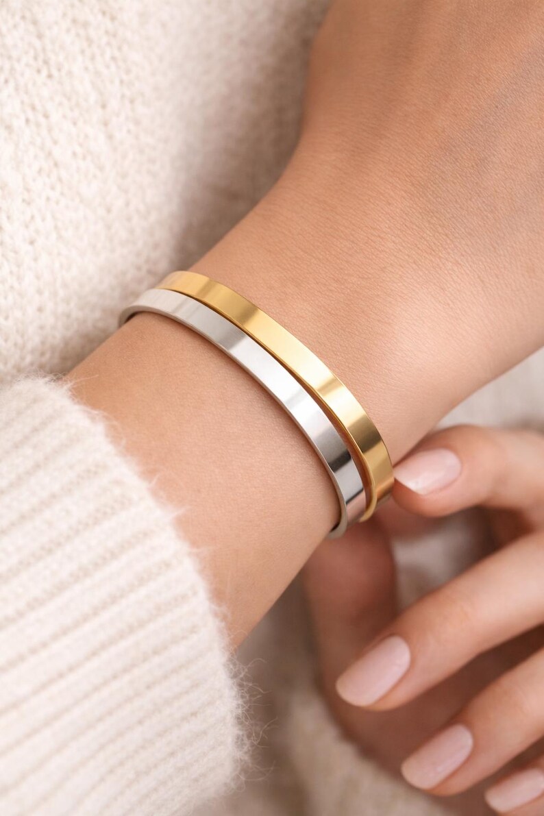 K&ouml;nnte beinhalten: Nahaufnahme von zwei minimalistischen Armb&auml;ndern, eines goldfarben und eines silberfarben. Die Armb&auml;nder werden am Handgelenk getragen, vor dem Hintergrund eines weichen, cremefarbenen Pullovers. Schlichtes, elegantes Design.