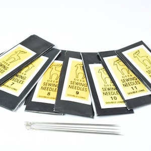 May include: A collection of seven black paper packets containing Deer Sewing Needles, labelled with sizes 6 through 12 and marked as double long. A group of silver metal sewing needles is arranged horizontally in front of the packets.