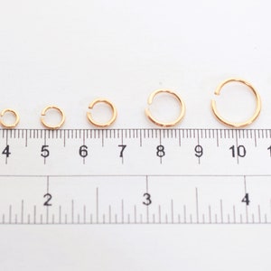 May include: A series of seven gold-colored open jump rings arranged in increasing size from left to right, positioned above a ruler showing measurements in both centimeters and inches to indicate their varying diameters.