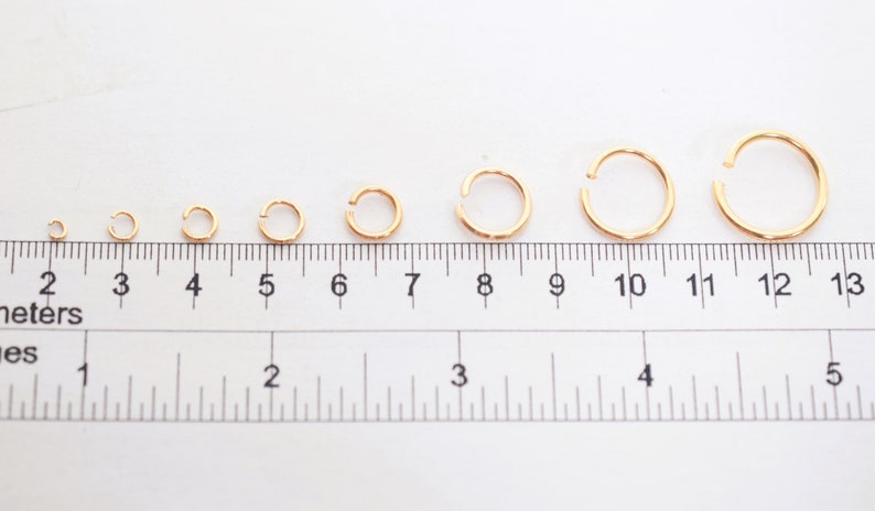 May include: A series of seven gold-colored open jump rings arranged in increasing size from left to right, positioned above a ruler showing measurements in both centimeters and inches to indicate their varying diameters.