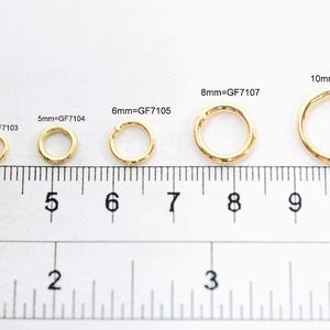 May include: A series of eight gold-toned metal jump rings arranged by size from 2mm to 12mm, positioned above a ruler showing both inches and centimeters. Each ring is labeled with its size and code, such as 2mm=GF7101 through 12mm=GF7110.