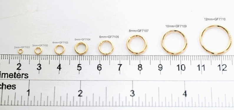 May include: A series of eight gold-toned metal jump rings arranged by size from 2mm to 12mm, positioned above a ruler showing both inches and centimeters. Each ring is labeled with its size and code, such as 2mm=GF7101 through 12mm=GF7110.