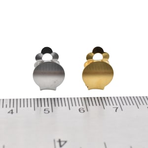 May include: Two metal earring backs, one silver and one gold, are shown next to a ruler. The backs are about 1 cm in diameter.