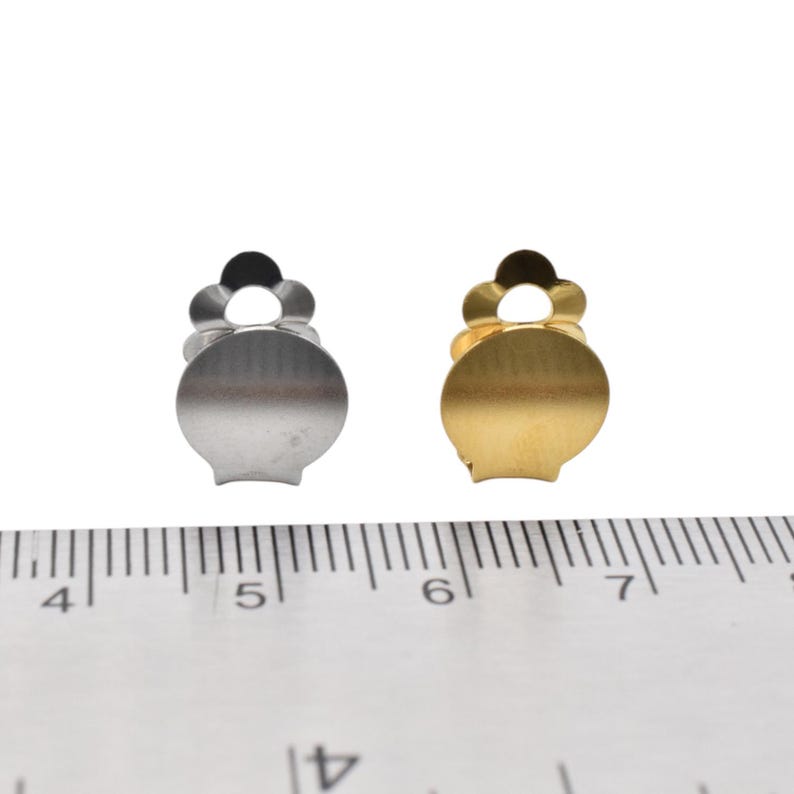 May include: Two metal earring backs, one silver and one gold, are shown next to a ruler. The backs are about 1 cm in diameter.