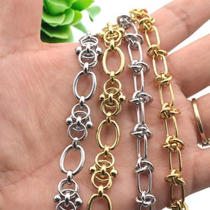 May include: Close-up of several metal chains in silver and gold tones. The chains have various designs, including oval links, decorative knots, and ornate connectors. The chains are displayed against a neutral background.