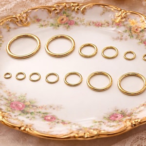May include: A collection of twelve smooth, polished gold-tone metal rings arranged in two rows on a decorative plate. The rings vary in diameter, ranging from small to large, and feature a simple, circular, seamless design with a reflective finish.