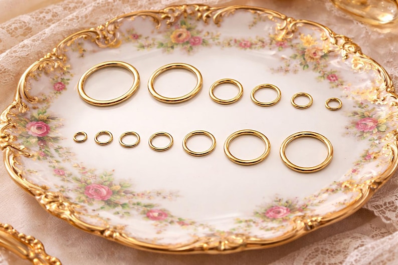 May include: A collection of twelve smooth, polished gold-tone metal rings arranged in two rows on a decorative plate. The rings vary in diameter, ranging from small to large, and feature a simple, circular, seamless design with a reflective finish.