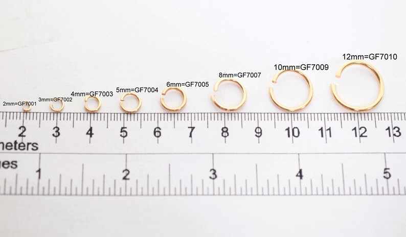 May include: A series of eight gold-colored metal jump rings arranged by size above a ruler. The rings are labeled with their diameters and product codes: 2mm=GF7001, 3mm=GF7002, 4mm=GF7003, 5mm=GF7004, 6mm=GF7005, 8mm=GF7007, 10mm=GF7009, and 12mm=GF7010.