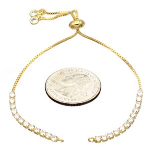 Puede incluir: Un brazalete de cadena dorada con un cierre deslizante y una fila de piedras de circonita c&uacute;bica transparentes. El brazalete se muestra junto a una moneda de 1 euro para referencia de tama&ntilde;o.