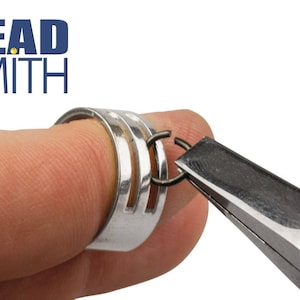 May include: A close-up of a silver ring with two openings, being used to hold a jump ring open with a pair of pliers. The ring is on a finger.