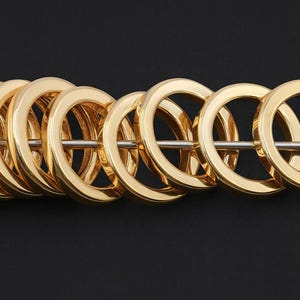 May include: A row of gold-colored, circular rings strung on a silver rod. The rings are evenly spaced and have a smooth, reflective surface. The background is a solid black.