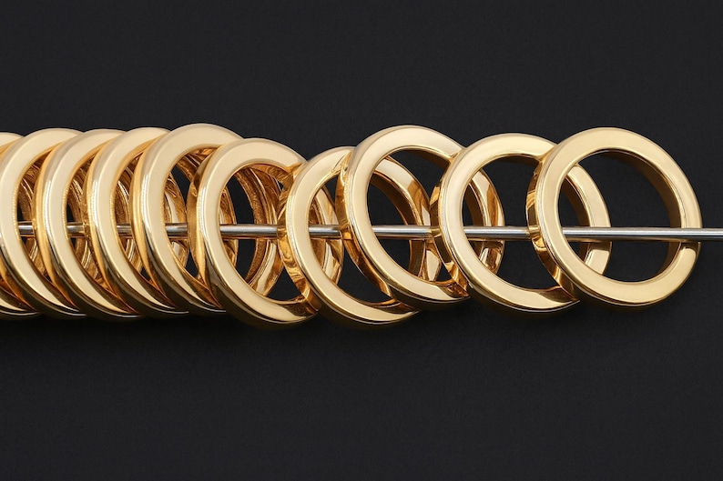 May include: A series of polished gold-tone metal rings arranged in a row, each featuring a smooth, rounded surface and a circular opening. The rings are threaded onto a thin, straight silver-colored metal rod that passes through their centers.