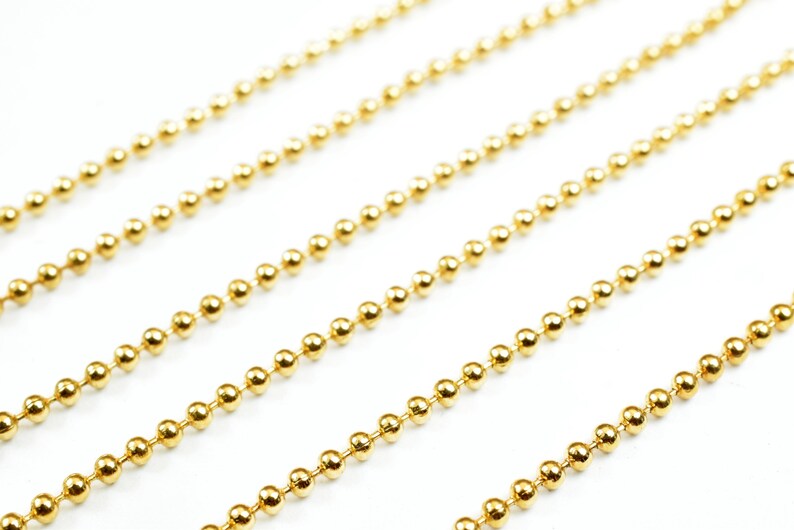 May include: Four strands of gold ball chain, a type of jewelry chain, are laid out in parallel lines on a white background.