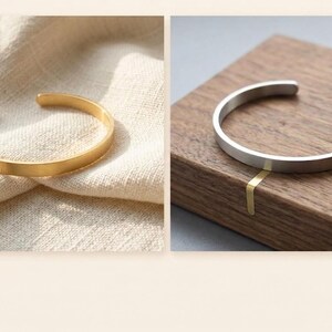 K&ouml;nnte beinhalten: Zwei offene Armreifen. Einer ist goldfarben und liegt auf einem beigen Stoff. Der andere ist silberfarben und wird auf einer Holzoberfl&auml;che pr&auml;sentiert. Beide Armreifen haben ein schlichtes, minimalistisches Design.
