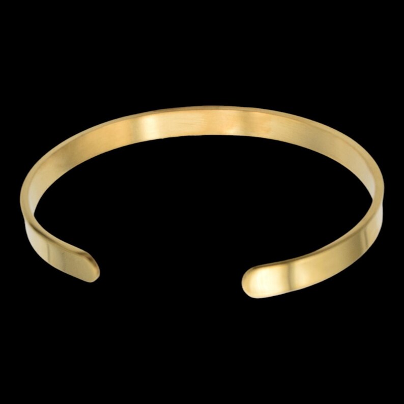 K&ouml;nnte beinhalten: Ein goldfarbenes Armreif-Armband mit einem einfachen, offenen Design. Das Armband hat eine glatte, polierte Oberfl&auml;che und eine leicht abgerundete Form. Die offenen Enden erm&ouml;glichen ein einfaches Tragen und Anpassen. Der Hintergrund ist schwarz.