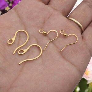 May include: Close-up of several gold-colored earring hooks. The hooks are in various styles, including French hooks and fish hooks. The hooks are made of a shiny, metallic material and are displayed against a neutral background.