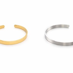 K&ouml;nnte beinhalten: Zwei offene Armreifen. Einer ist goldfarben, der andere silberfarben. Beide Armreifen haben ein schlichtes, minimalistisches Design. Die Armreifen sind aus Metall und werden auf wei&szlig;em Hintergrund gezeigt.