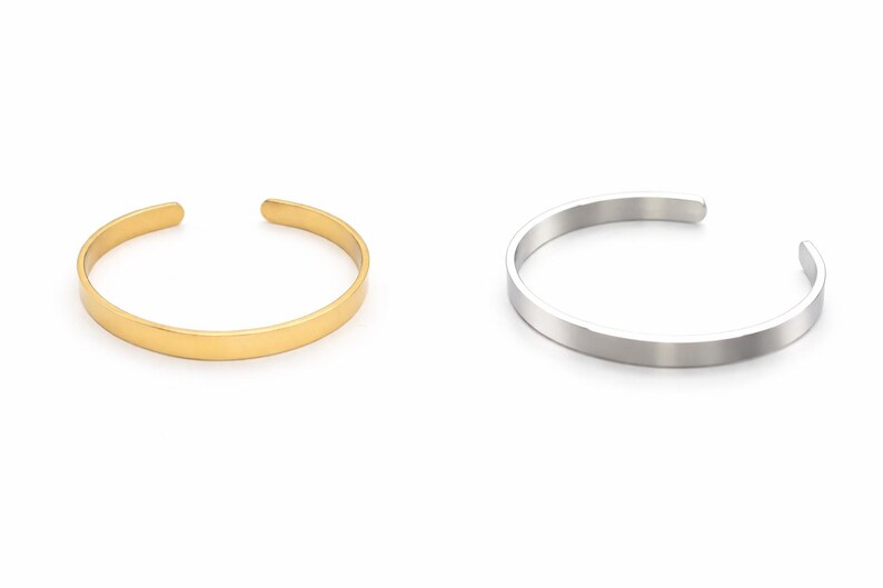 K&ouml;nnte beinhalten: Zwei offene Armreifen. Einer ist goldfarben, der andere silberfarben. Beide Armreifen haben ein schlichtes, minimalistisches Design. Die Armreifen sind aus Metall und werden auf wei&szlig;em Hintergrund gezeigt.