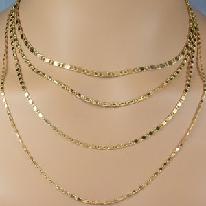 May include: Five gold chains of varying lengths, layered on a model's neck. The chains are made of small, flat, rectangular links.