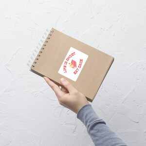 Könnte beinhalten: Ein weißer Aufkleber mit dem Text "Life is short eat cake" in Rot, mit einer Kuchengrafik. Der Aufkleber befindet sich auf einem braunen Spiralnotizbuch. Das Notizbuch wird von einer Person gehalten.