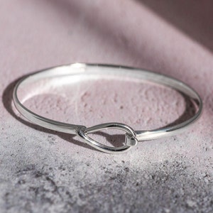 Op de afbeelding: Een zilveren armband met een simpel, elegant ontwerp. De armband heeft een gladde, gepolijste afwerking en een unieke sluiting in de vorm van een traan.