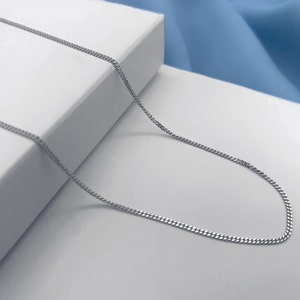 May include: A silver chain necklace with a delicate curb link design. The chain is laying on a white surface.