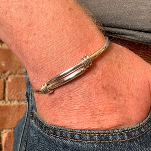Könnte beinhalten: Ein silbernes Armreif-Armband mit einem einfachen, geknoteten Design. Das Armband wird am Handgelenk getragen.