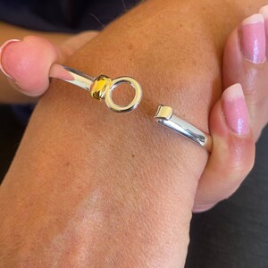 Puede incluir: Una pulsera de puño plateada con un acento dorado en el cierre. La pulsera está abierta, mostrando el mecanismo de cierre. La pulsera está en una muñeca.