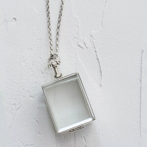 Könnte beinhalten: Eine silberne Halskette mit einem Anhänger aus klarem Glas. Der Anhänger hängt vor einem weißen Hintergrund.