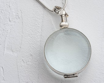 May include: A silver-toned magnifying glass pendant suspended from a matching snake chain. The circular lens is encased in a polished metal frame with a decorative screw-top bail, featuring a clear glass surface and a hinged bottom closure.