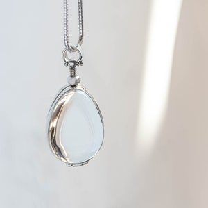 Puede incluir: Un collar con colgante plateado con un relicario en forma de lágrima. El relicario tiene una superficie clara y reflectante y está suspendido de una delicada cadena. El fondo es de un color suave y neutro, con una sutil sombra.