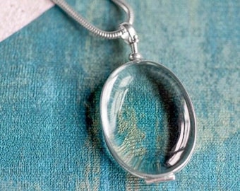May include: A silver chain necklace with a round magnifying glass pendant. The pendant is made of metal and glass and has a clear lens.