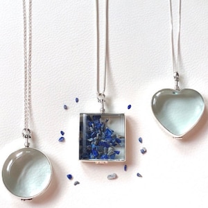 Op de afbeelding: Drie zilveren kettingen met doorzichtige glazen hangers. Eén hanger is rond, één is vierkant en gevuld met blauwe stenen, en één is hartvormig. De kettingen worden gepresenteerd op een witte ondergrond.
