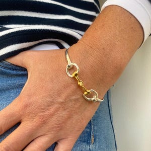 Könnte beinhalten: Silber- und goldfarbenes Armband mit einem Gebiss-Design. Das Armband hat ein Scharnierdesign mit einem goldfarbenen Mittelstück und silberfarbenen Ringen. Das Armband wird an einem Handgelenk mit einem blauen Jeanshemd und einem gestreiften Hemd getragen.