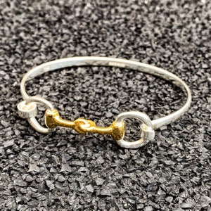 May include: A silver bangle bracelet with a gold-toned horse bit charm. The bracelet is open and has a simple design.
