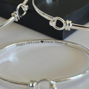 Puede incluir: Pulseras de brazalete plateadas con cierre de gancho. Una pulsera está enfocada, mostrando el texto grabado: "Happy 18th Lexie ❤️ forever Nanna xx". Las pulseras están sobre una superficie clara con un fondo oscuro.