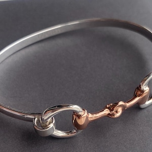 May include: A silver bangle bracelet with a rose gold horse bit design. The bracelet is open and has a simple, elegant design.