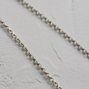 May include: A close-up of two silver chains on a white textured background. The chains are made of small, linked circles.