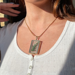 May include: A gold chain necklace with a rectangular pendant featuring a pressed green leaf inside a clear glass frame.