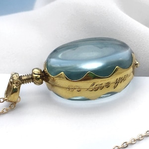 Op de afbeelding: Een gouden ketting met een blauwe glazen ovale hanger. De hanger heeft een gegolfde rand en de inscriptie "we love you" is gegraveerd in het glas.
