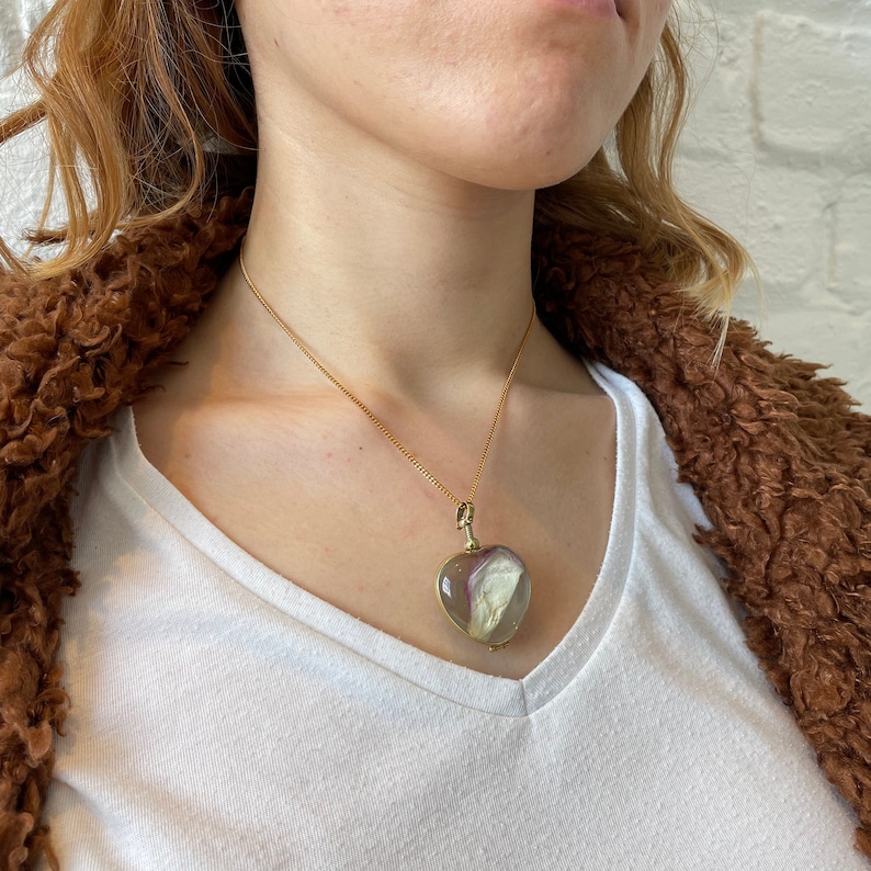 May include: A gold chain necklace with a heart-shaped locket pendant. The locket is clear and contains a dried flower.